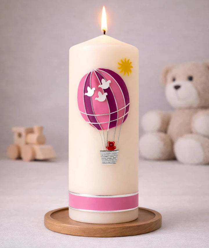 elfenbeinfarbene Taufkerze mit rosa Heißluftballon und kleinem Teddy im Korb, brennend auf Holzsockel vor neutralem Hintergrund mit Teddybär