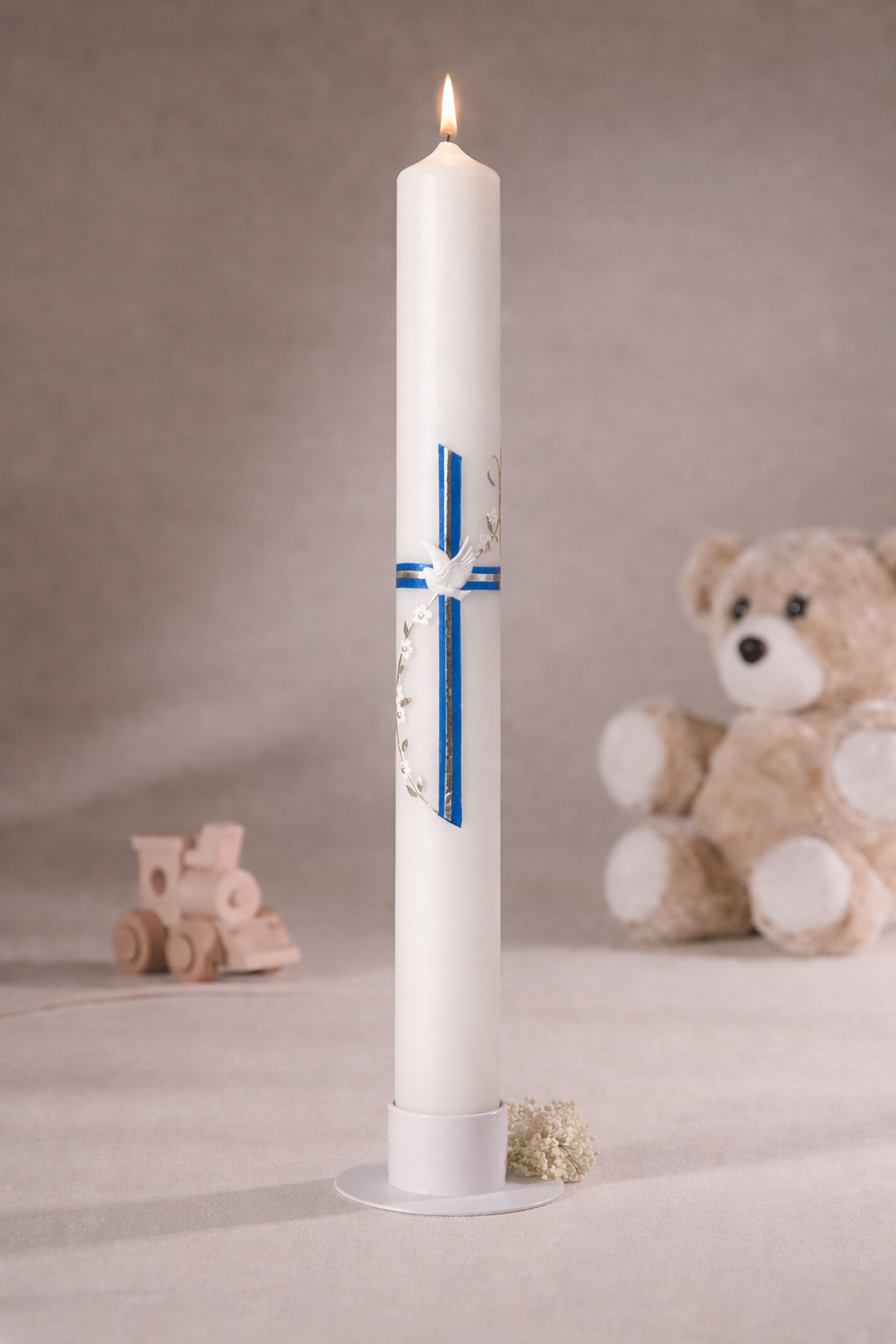 Weiße Taufkerze mit blauem Kreuz und weißer Friedenstaube, stehend auf Kerzenteller vor neutralem Hintergrund mit Teddybär und Holzspielzeug
