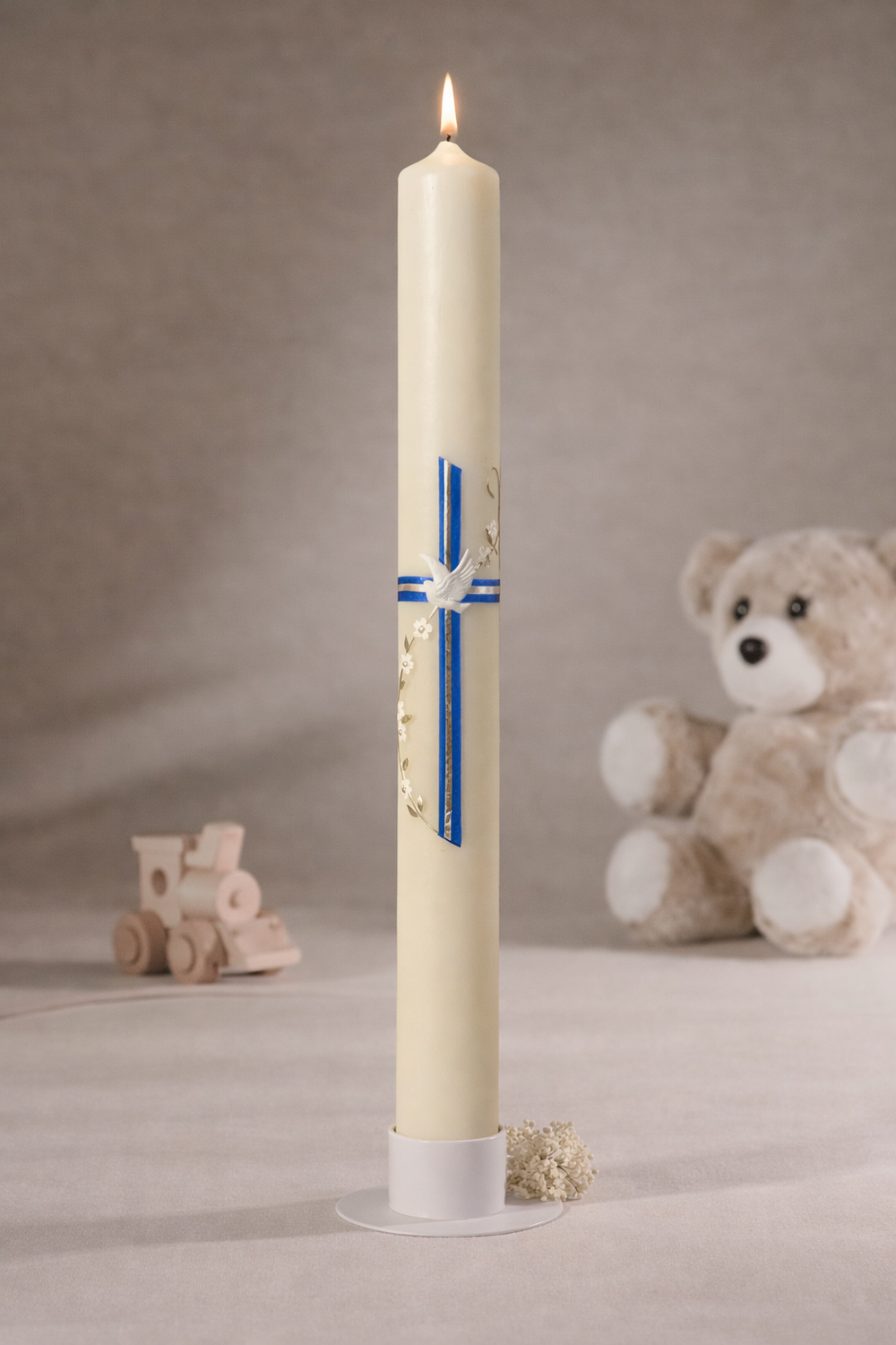 Weiße Taufkerze mit blauem Kreuz und goldfarbenen Verzierungen, brennend auf Kerzenteller vor neutralem Hintergrund mit Teddybär und Holzspielzeug