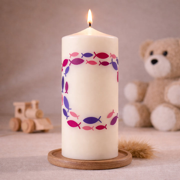 Elfenbeifarbige Taufkerze mit Ichthys-Fischsymbolen im Kreis in Rosa und Lila, stehend auf Holzsockel vor neutralem Hintergrund mit Teddybär
