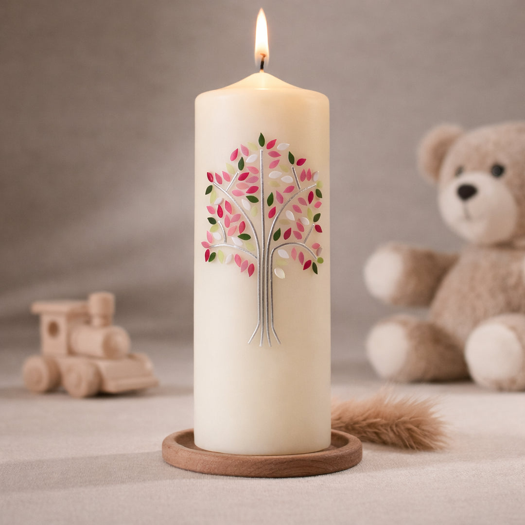 Weiße Taufkerze mit Lebensbaum-Motiv in Rosa, Pink und Grün, brennend auf Holzsockel vor neutralem Hintergrund mit Teddybär