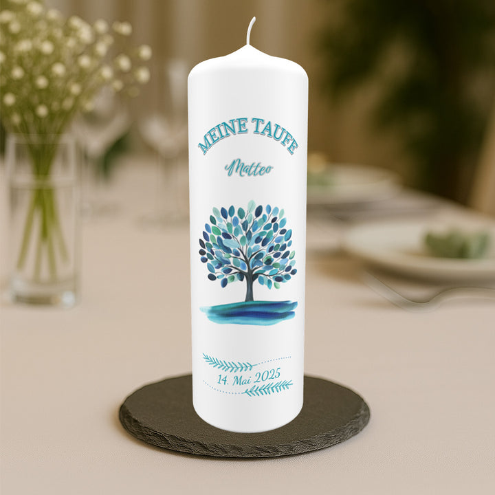 Weiße Taufkerze mit blau-grünem Baum-Motiv, dem Schriftzug „Meine Taufe“, dem Namen „Matteo“ und dem Taufdatum „14. Mai 2025“, arrangiert auf einem gedeckten Tisch mit unscharfem Hintergrund.