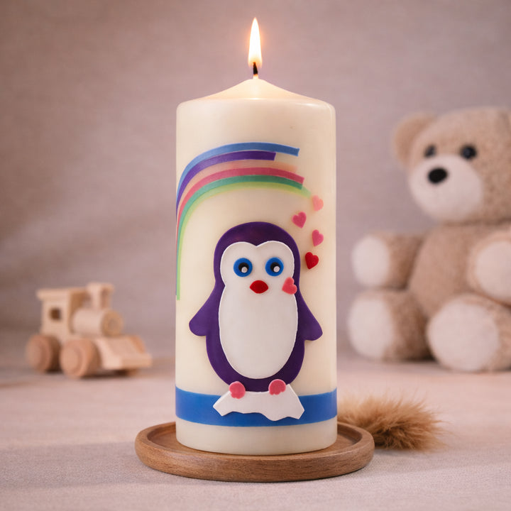 Elfenbein Taufkerze mit Pinguin und Regenbogenmotiv, stehend auf Holzsockel vor neutralem Hintergrund mit Teddybär
