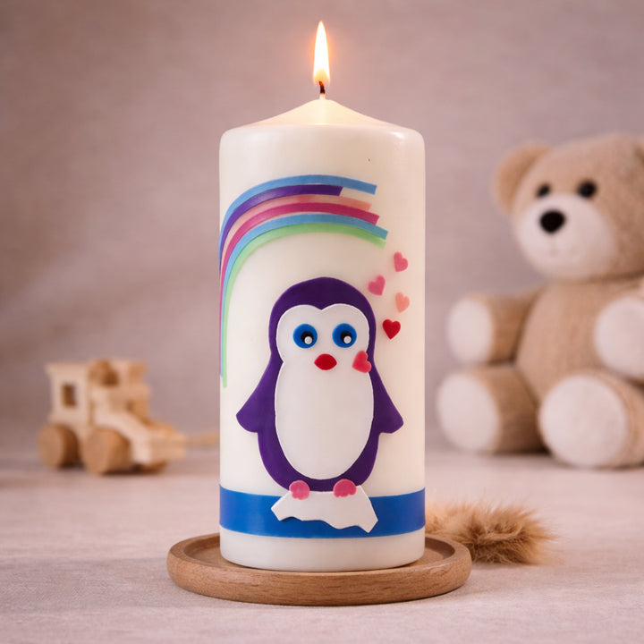 Weiße Taufkerze mit Pinguinmotiv unter buntem Regenbogen und kleinen Herzsymbolen, brennend auf Holzsockel vor neutralem Hintergrund mit Teddybär