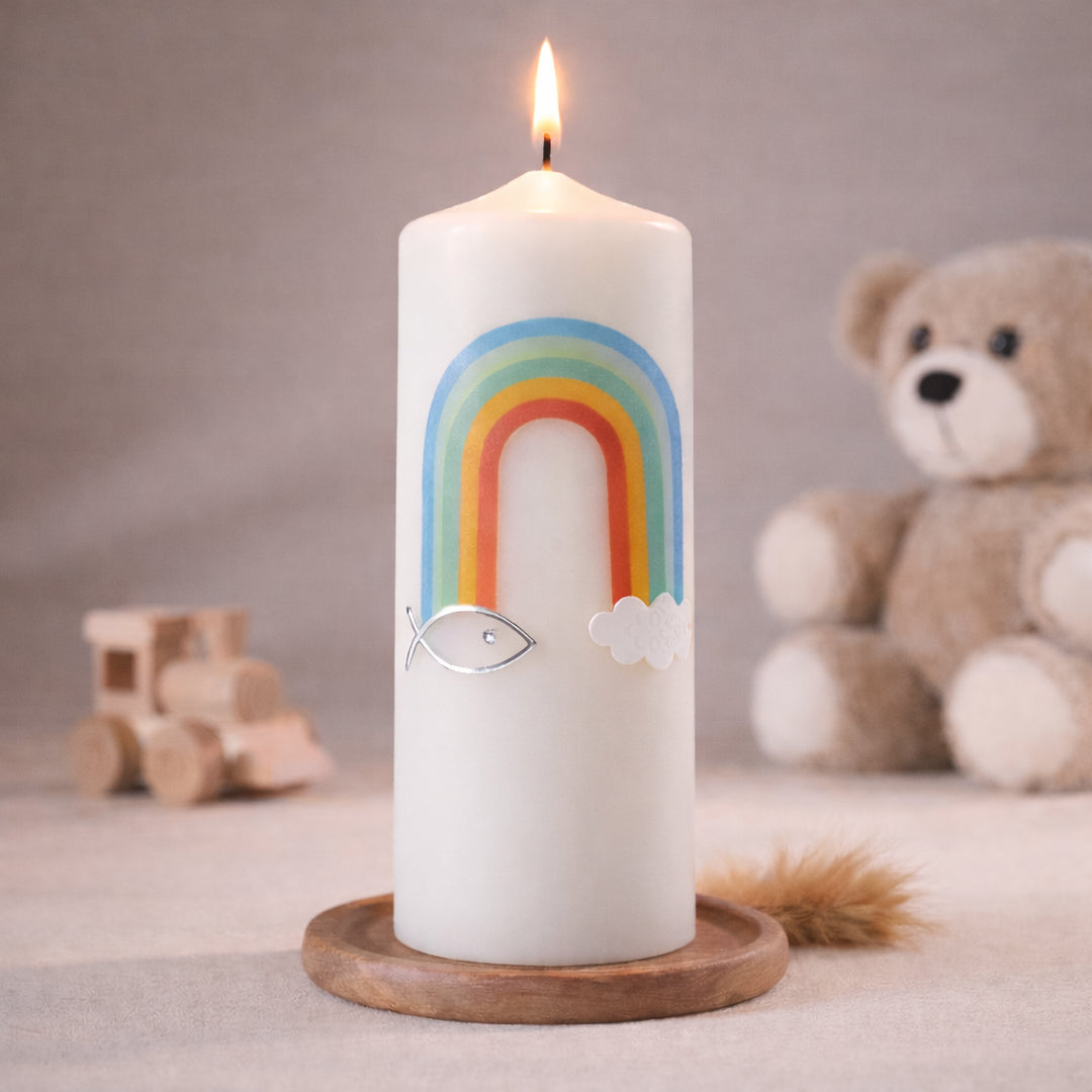 Weiße Taufkerze mit buntem Regenbogen, Ichthys-Fischsymbol und Wolkenmotiv, brennend auf Holzsockel vor neutralem Hintergrund mit Teddybär