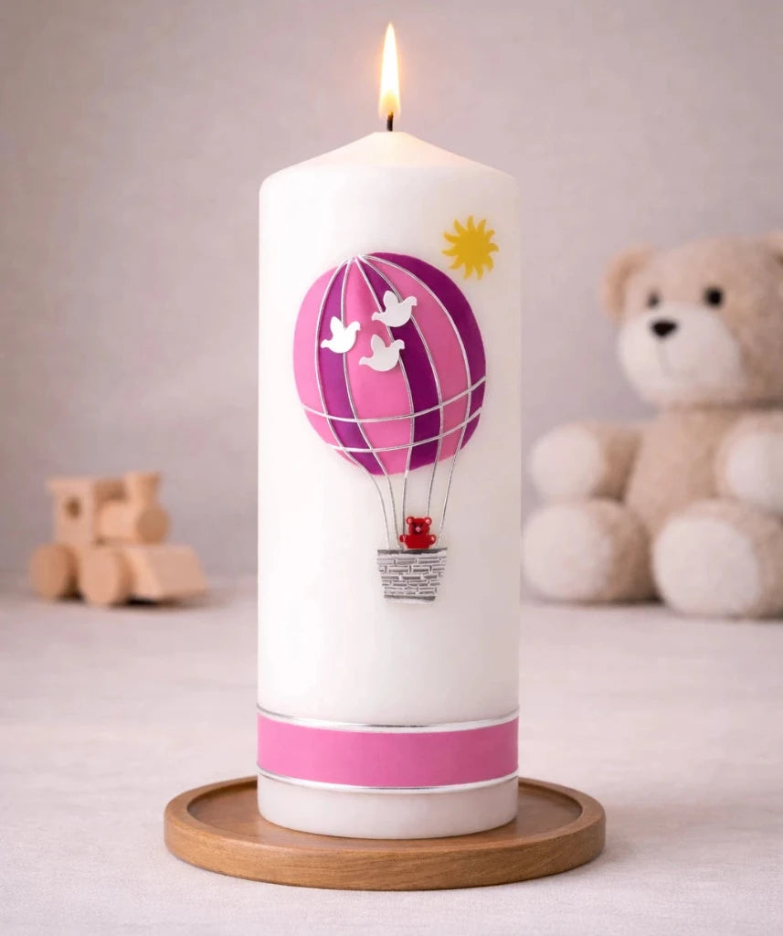 Weiße Taufkerze mit rosa Heißluftballon und kleinem Teddy im Korb, brennend auf Holzsockel vor neutralem Hintergrund mit Teddybär