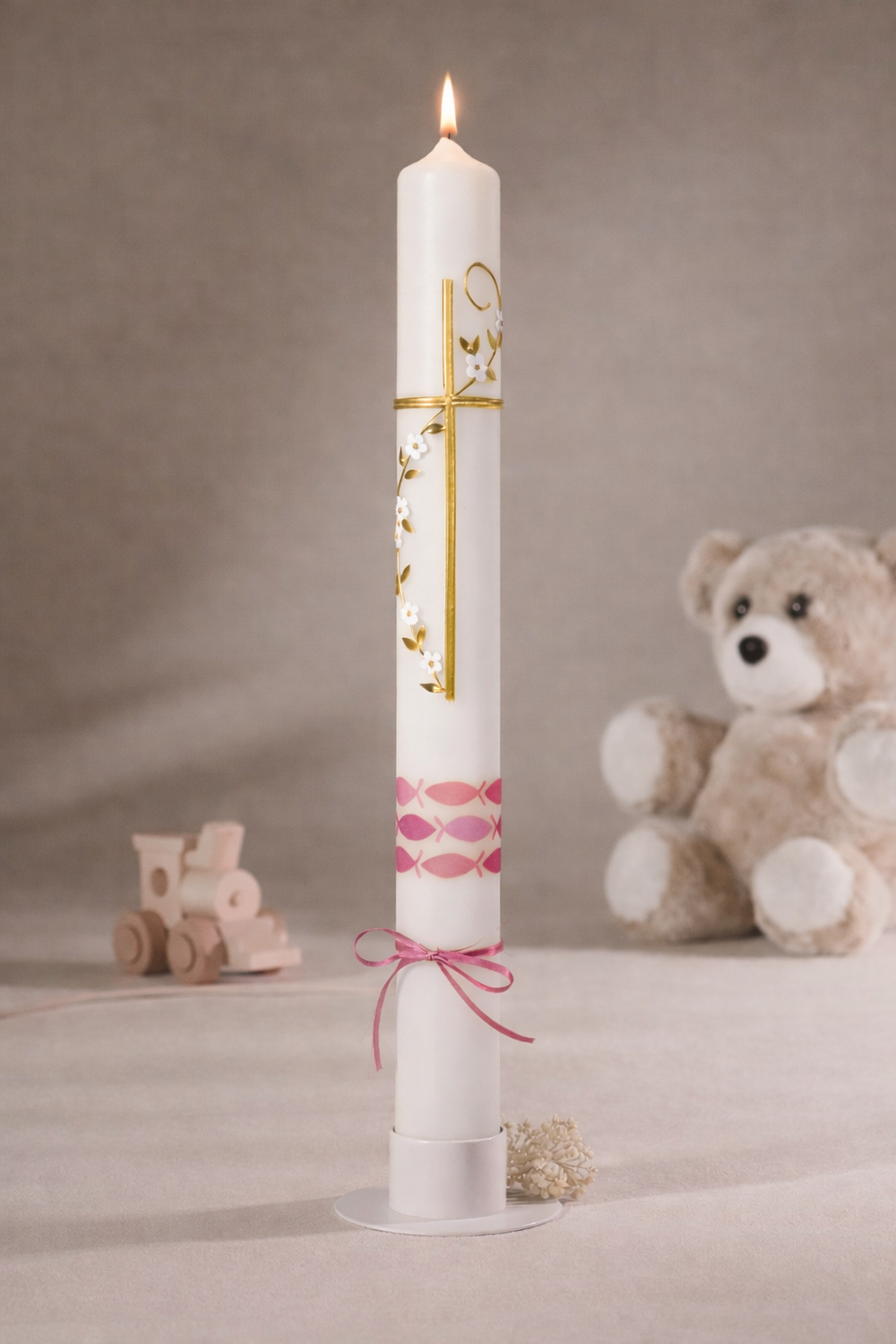 Taufkerze mit rosa Fischmotiv und Goldkreuz in seitlicher Ansicht, dekoriert mit rosa Band, fotografiert vor neutralem Hintergrund mit Teddybär und Holzspielzeug
