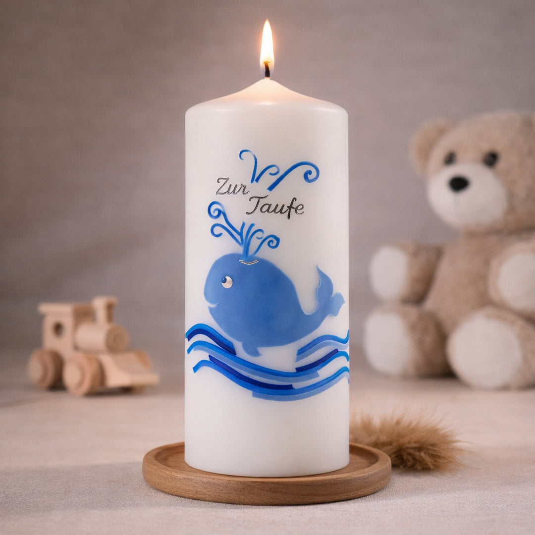 Weiße Taufkerze mit blauem Wal und Wellenmotiv, brennend auf Holzsockel vor neutralem Hintergrund mit Teddybär