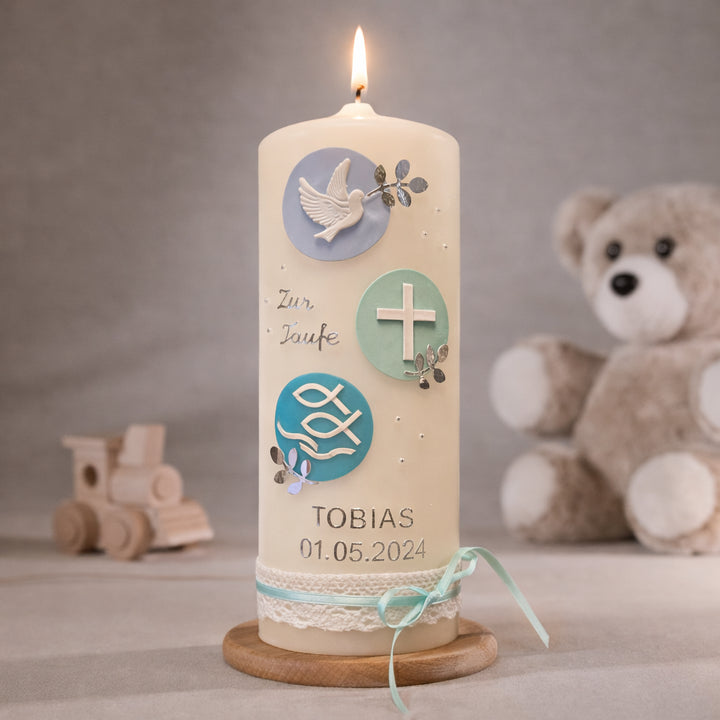 Cremefarbene Taufkerze mit runden Zierelementen in Mint und Blau, Taube, Kreuz und Fischsymbol sowie personalisiertem Namen und Datum, brennend auf Holzsockel vor neutralem Hintergrund mit Teddybär
