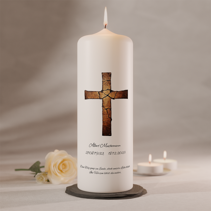 Weiße Trauerkerze mit gebrochenem Kreuz in Kupferoptik und Namensbeschriftung, stehend auf dunklem Kerzenteller neben weißer Rose und zwei Teelichtern