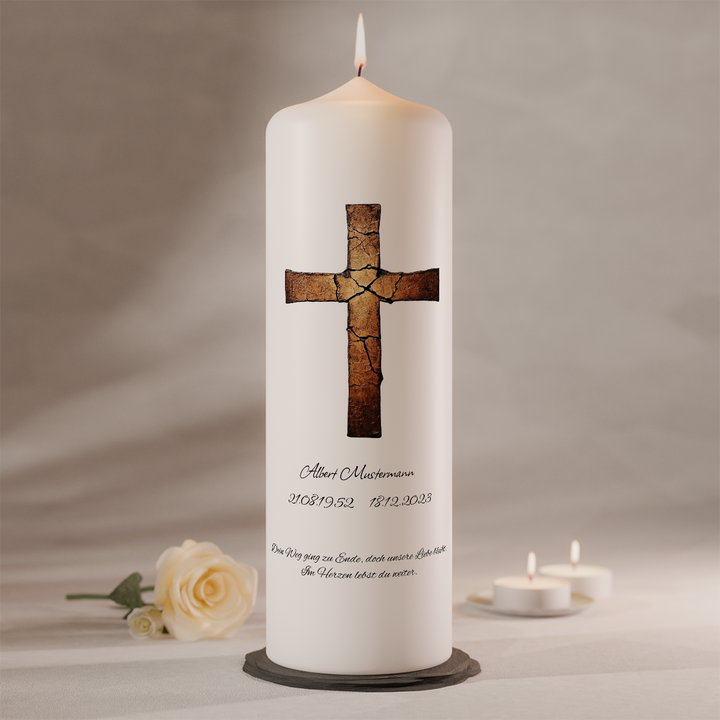 Weiße Trauerkerze mit gebrochenem Kreuz in Kupferoptik und individueller Beschriftung, brennend auf dunklem Kerzenteller neben weißer Rose und Teelichtern