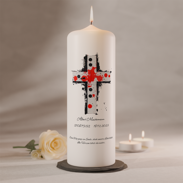 Weiße Trauerkerze mit künstlerischem Kreuz in Rot und Schwarz und individueller Namensbeschriftung, brennend auf dunklem Kerzenteller neben weißer Rose und zwei Teelichtern
