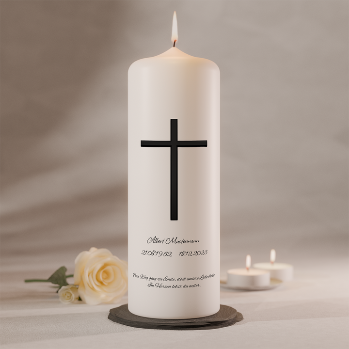 Weiße Trauerkerze mit schwarzem Langkreuz und Namensbeschriftung, brennend auf dunklem Kerzenteller neben weißer Rose und zwei Teelichtern