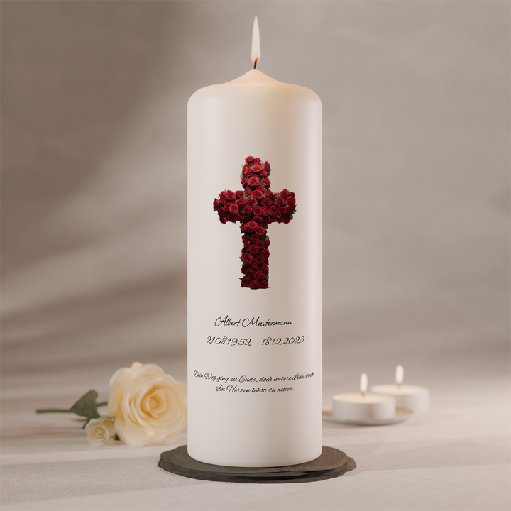 Weiße Trauerkerze mit rotem Rosenkreuz und Namensbeschriftung, brennend auf dunklem Kerzenteller neben weißer Rose und zwei Teelichtern