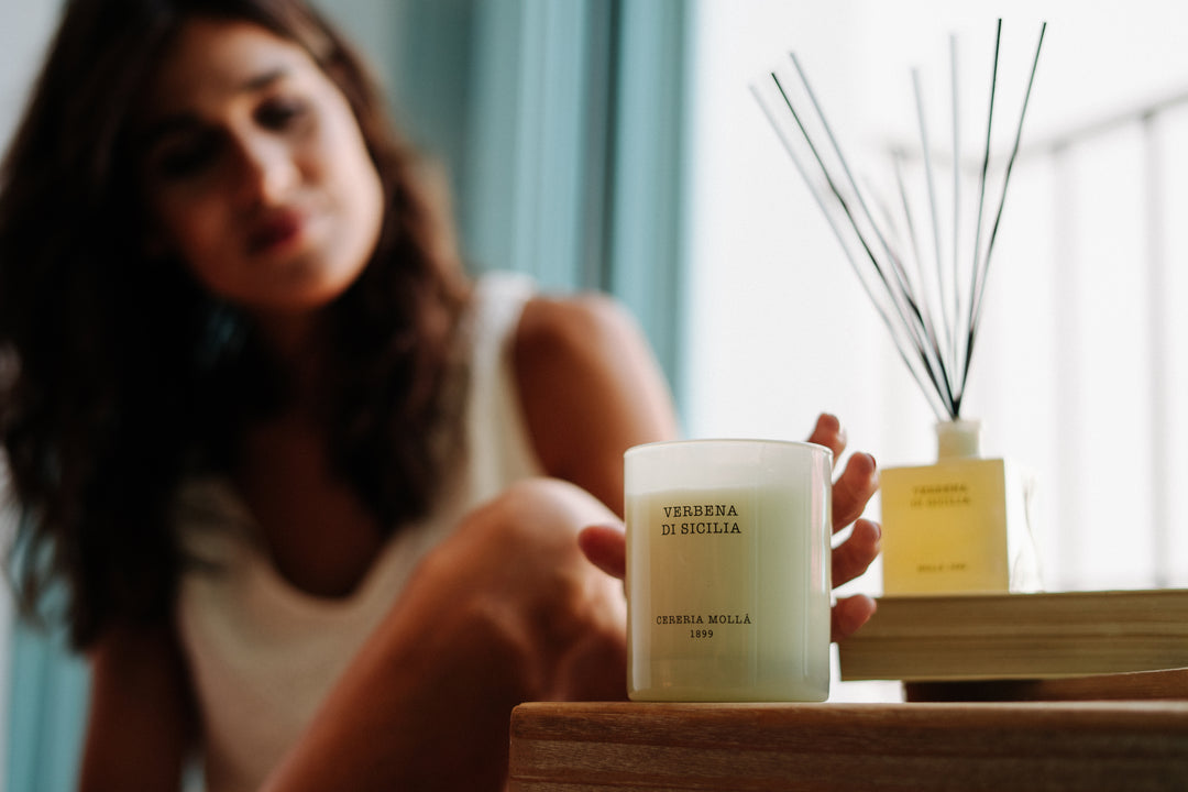 Verbena di Sicilia Duftkerze von Cereria Mollá 1899, auf Holztablett mit Frau im Hintergrund, sanftes Tageslicht, Duft von Zitronenverbene und mediterranen Kräutern.