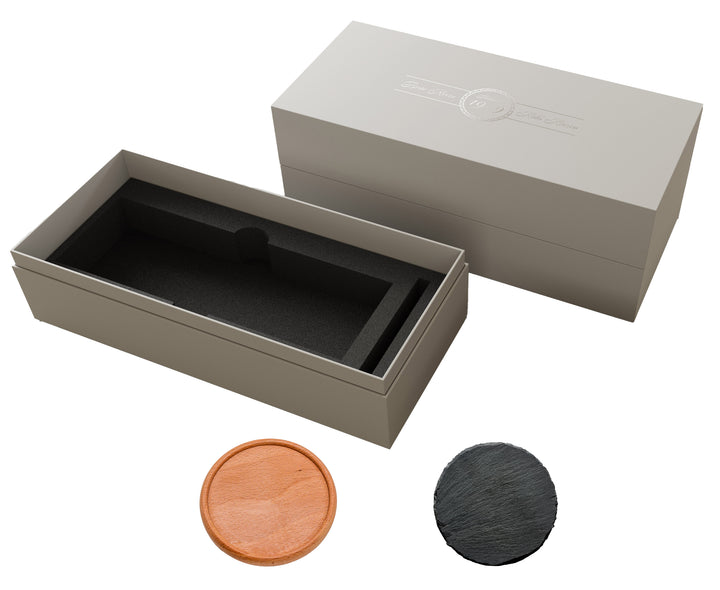 Graue Geschenkbox mit Deckel und schwarzer Schaumstoffeinlage, daneben zwei runde Untersetzer – einer aus Holz, einer aus Schiefer – auf weißem Hintergrund